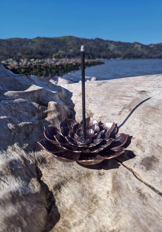 Lotus shaped incense holder, sitting on log, single stick of burning incense, water and blue sky in the backdrop
