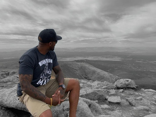 Tatooed African American male wearing brown shorts, grey shirt, and black hat with full sleeve tattoos. Sitting on a rock overlooking a valley.