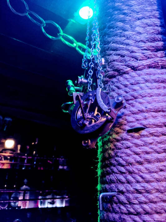 Sextant hanging from rope covered post, green light shining on sextant, dark lighting in background shining on rum bottles