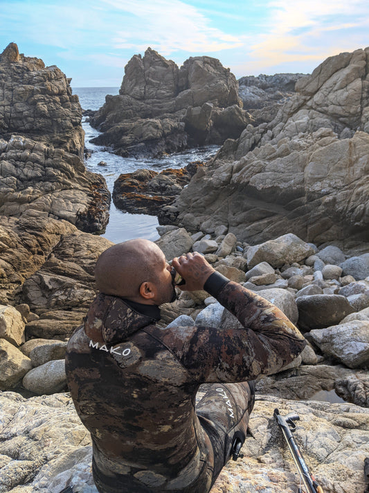 black male sitting on rocky shoreline, wearing wetsuit, drinking a beer,