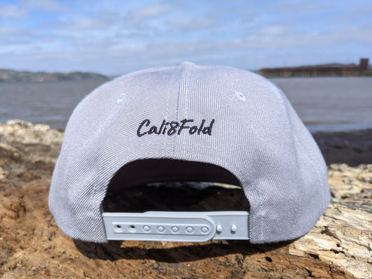 Grey hat, cali8fold written on back, hat sitting on log in front of cove of water