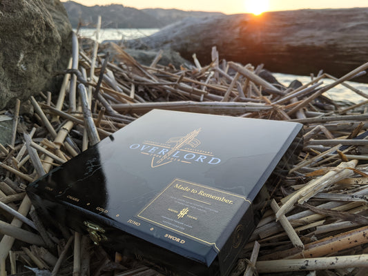 Black box of cigars, overlord written on box, box is laying on top of drift wood, sun setting in background