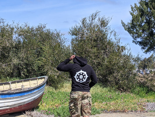 Man wearing black hoodie, buddhist eightfold path wheel on back, camo shorts, standing next to boat, trees in background,
