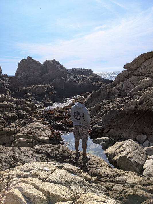 Man standing at rocky coast, wearing a grey sweater, black Buddhist eightfold symbol on back, camo pants, blue skies, small pool of water in front, cali8fold