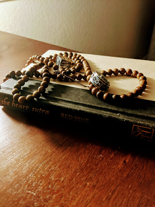 "Heart Sutra" book on desk, warm lighting, prayer beads on top of book, two silver rings laying on beads, book has black spine and beige cover, "Heart Sutra" written in gold lettering on spine. Cali8Fold
