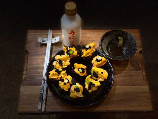 Prepared purple sea urchin rolls, ten on a black plate, on a wooden platter, white chopsticks next to it, saucer with black liquid on upper right, white bottle with Japanese characters above plate